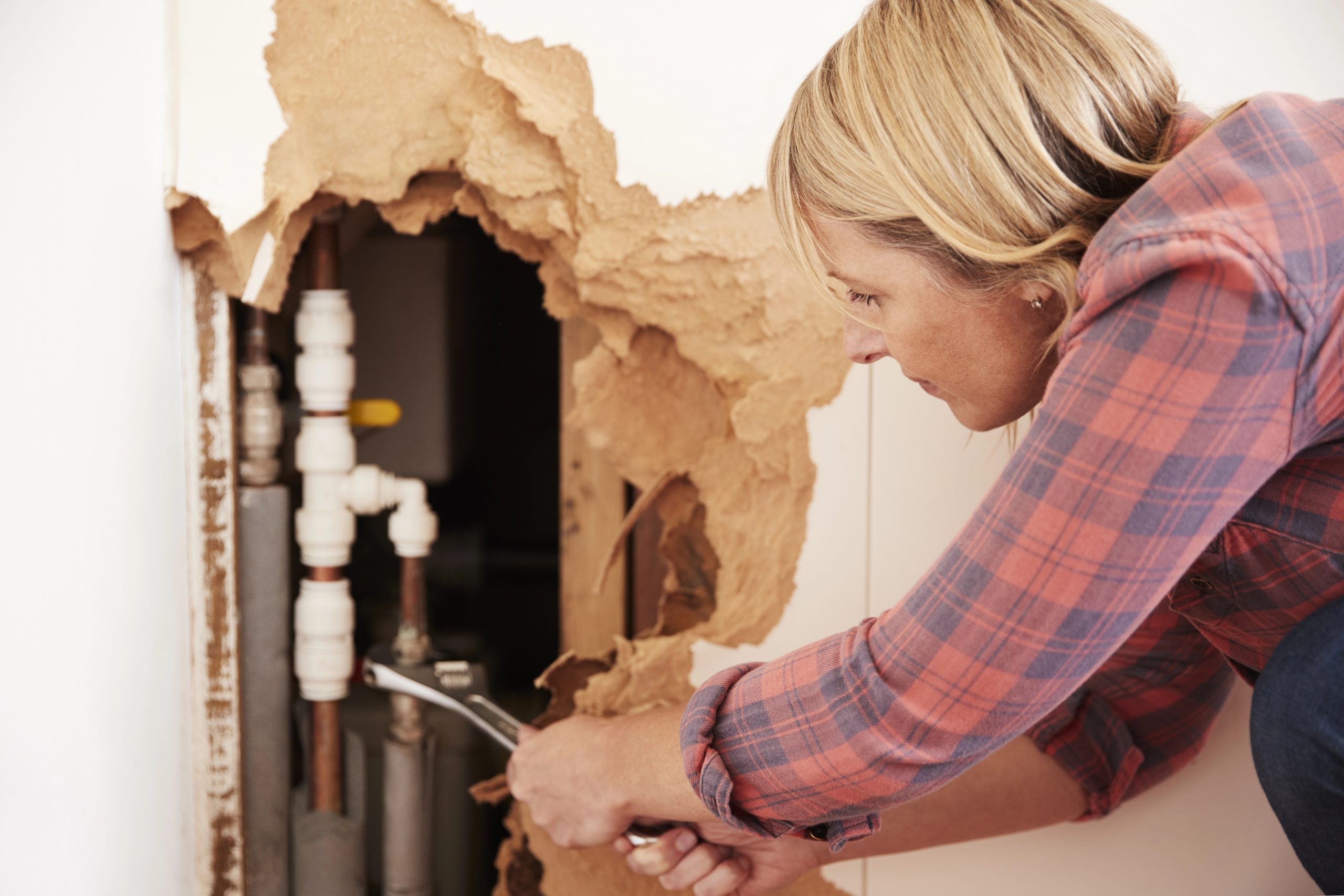 Woman using a wrench to repair exposed water pipes inside a wall after severe water damage, addressing plumbing issues to avoid future problems.