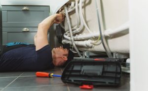 Emergency Plumber Brisbane: Professional plumber lying on the floor repairing pipes under a bathtub, with tools and a toolbox nearby.