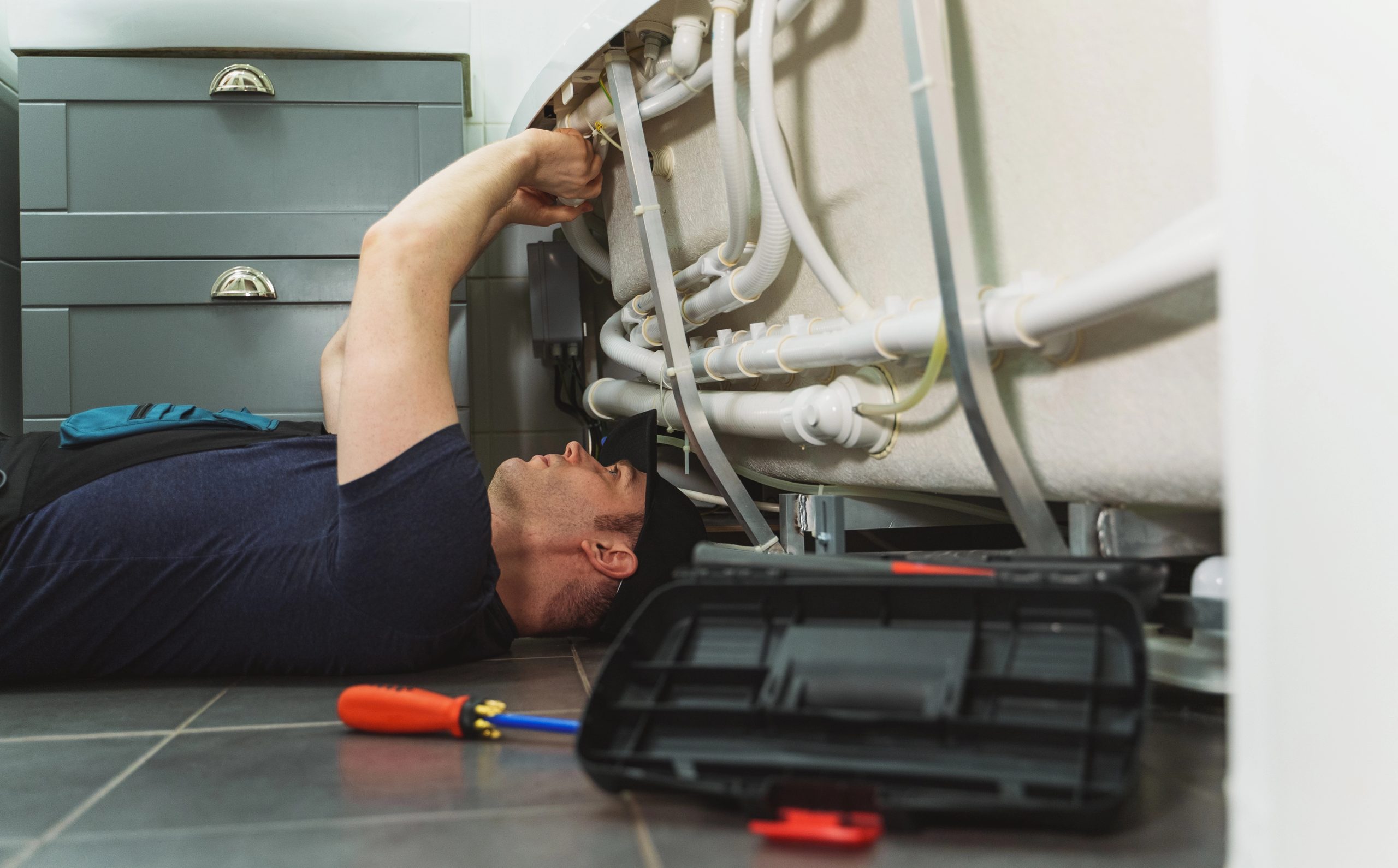 Emergency Plumber Brisbane: Professional plumber lying on the floor repairing pipes under a bathtub, with tools and a toolbox nearby.